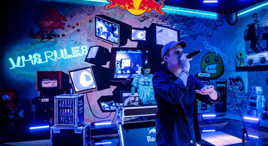 Jan rapowanie koncert w neonowym świetle. Na ścianie napisy "VHS RULES", logo Red Bulla i stare monitory.