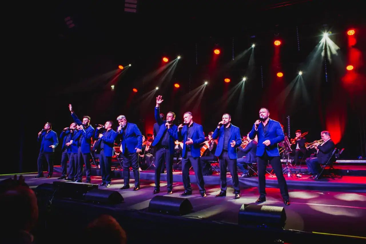 Dziesięciu tenorów w niebieskich marynarkach śpiewa na scenie, w tle orkiestra.