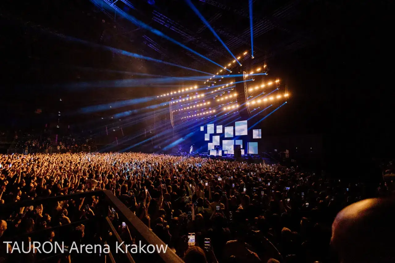 Tłum ludzi na koncercie rapowania, oświetlony sceną i światłami. Jan rapowanie koncert w TAURON Arenie Kraków.