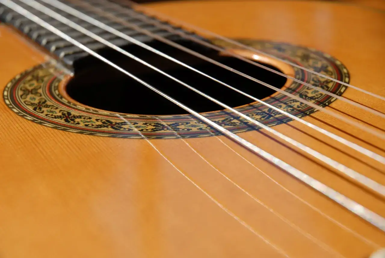 Zbliżenie na gitarę klasyczną. Widać jej drewniany korpus, ozdobną rozetę wokół otworu rezonansowego i sześć białych strun.