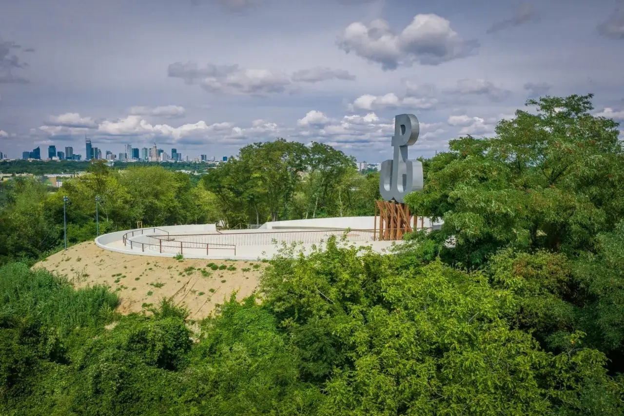 Symbol Polski Walczącej w parku akcji burza, na tle warszawskiej panoramy.