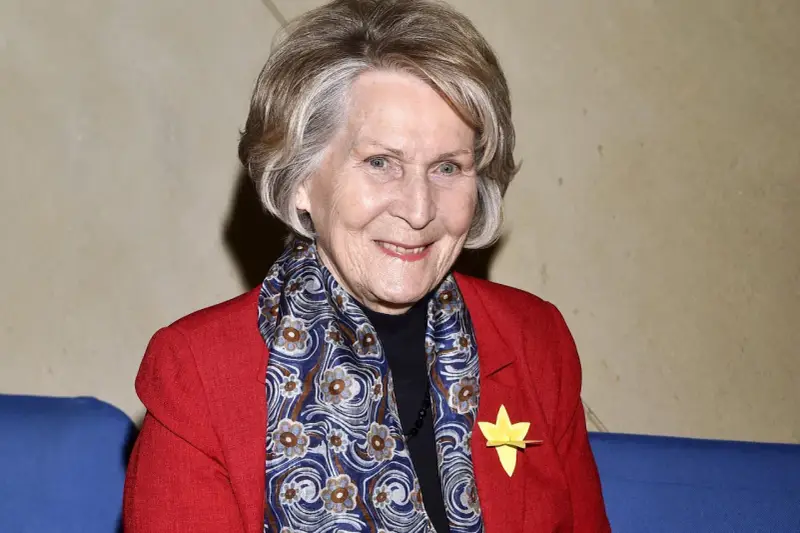Uśmiechnięta Irena Santor w czerwonej marynarce i wzorzystym szalu, z żółtą rozetką na klapie.